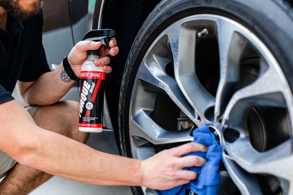 A person cleans a car wheel using a spray bottle and a cloth, ensuring a polished finish.
