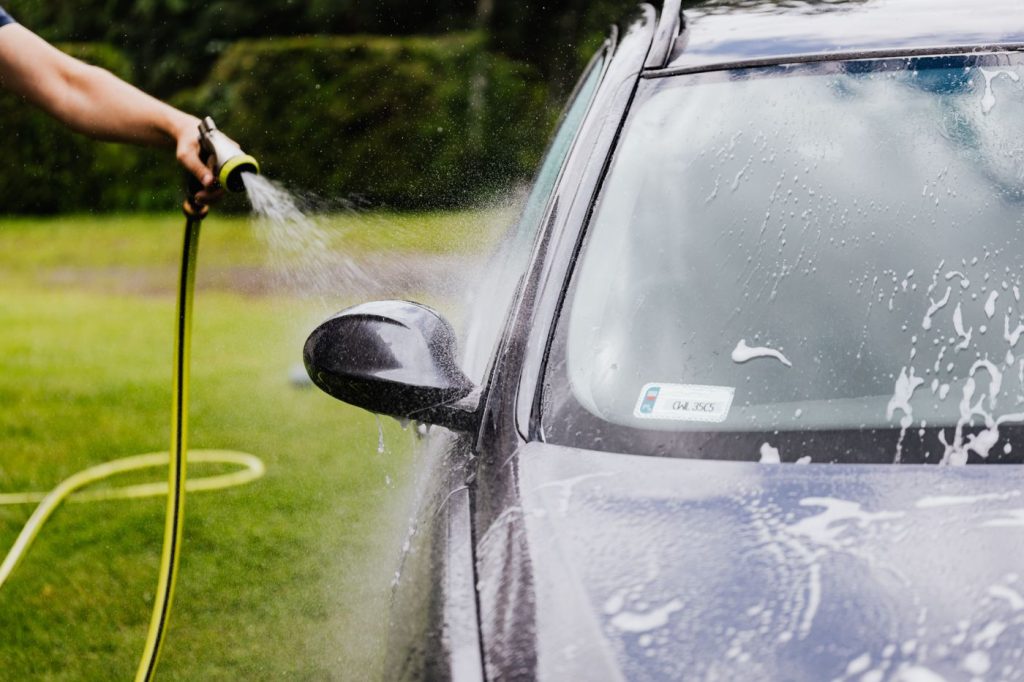 Person washing a car with a hose outdoors, creating suds on a sunny day.